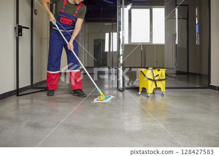 Janitor in uniform mopping floor inside modern commercial building during routine cleaning shift. Janitor in uniform mopping floor inside modern commercial building during routine cleaning shift. 128427583
