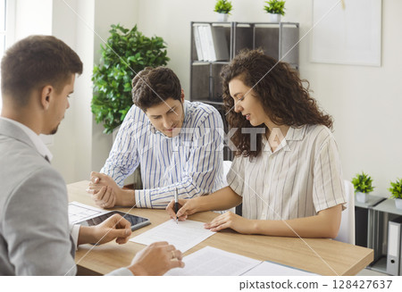 Young couple signing a contract with a man realtor or insurance agent sitting in office. 128427637