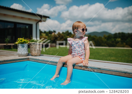 Little girl sitting by pool, splashing with legs in water. 128428203