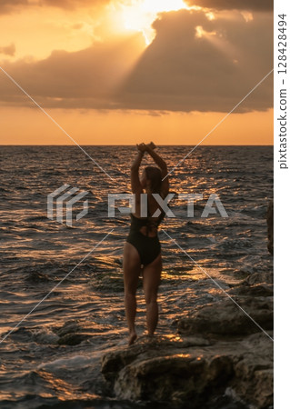 Woman Sunset Beach Pose - A woman in a black swimsuit poses on a rocky beach with the sunset in the background. Woman Sunset Beach Pose - A woman in a black swimsuit poses on a rocky beach with the sunset in the background. 128428494