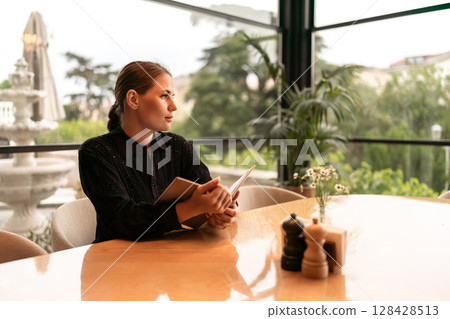 Woman Restaurant Menu: Woman reads menu in restaurant, daydreaming near the fountain, during daylight hours. Woman Restaurant Menu: Woman reads menu in restaurant, daydreaming near the fountain, during daylight hours. 128428513