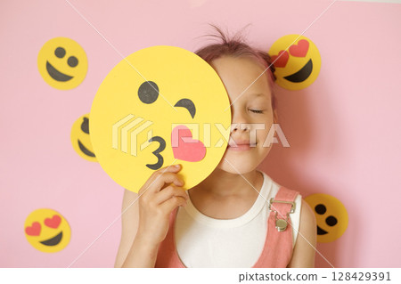 A cute little child on a pink studio background holds a smile face depicting a kiss. A girl surrounded by a variety of emoticons. World Emoji Day on July 17th A cute little child on a pink studio background holds a smile face depicting a kiss. A girl surrounded by a variety of emoticons. World Emoji Day on July 17th 128429391