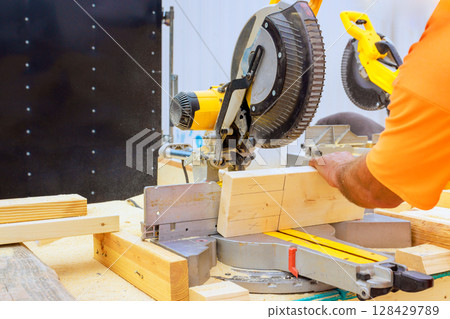 Skilled craftsman uses miter saw to precisely cut wooden boards in workshop filled with tools. Skilled craftsman uses miter saw to precisely cut wooden boards in workshop filled with tools. 128429789