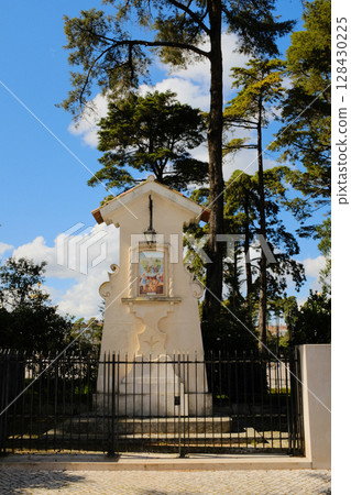 Catholic roadside shrine in Fatima Portugal with tiled Virgin Mary image and metal fence surrounded by pine trees under clear blue sky religious monument and local devotion site. High quality photo 128430225