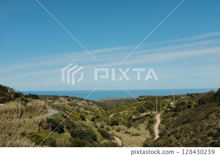 Scenic coastal road with view of Cabo da Roca lighthouse in Sintra Portugal curving highway through natural cliffs overlooking Atlantic Ocean under clear blue sky Scenic coastal road with view of Cabo da Roca lighthouse in Sintra Portugal curving highway through natural cliffs overlooking Atlantic Ocean under clear blue sky 128430239
