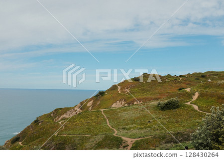 Hikers walking along scenic coastal trail at Cabo da Roca Sintra Portugal with grassy cliffs overlooking Atlantic Ocean under cloudy sky westernmost mainland Europe outdoor travel landscape. High 128430264