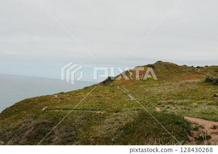 Hikers walking along scenic coastal trail at Cabo da Roca Sintra Portugal with grassy cliffs overlooking Atlantic Ocean under cloudy sky westernmost mainland Europe outdoor travel landscape. High 128430268