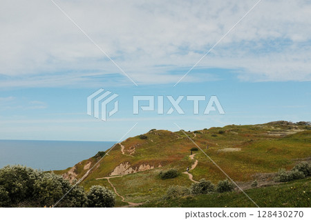 Hikers walking along scenic coastal trail at Cabo da Roca Sintra Portugal with grassy cliffs overlooking Atlantic Ocean under cloudy sky westernmost mainland Europe outdoor travel landscape. High 128430270