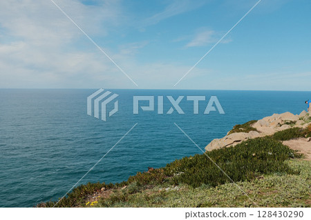 Scenic view of Cabo da Roca steep rocky cliffs meeting blue Atlantic Ocean under bright sky in Sintra Portugal panoramic coastal landscape from westernmost point of mainland Europe 128430290