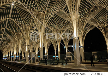 Modern illuminated architecture of Oriente Station at night in Lisbon Portugal featuring geometric roof design arches and empty railway tracks in futuristic urban environment. High quality photo 128430315