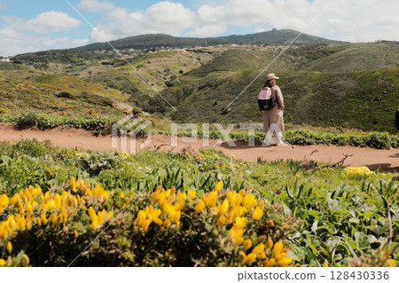 Woman enjoying springtime flower field on scenic hiking trail with mountain hills in background at Cabo da Roca Sintra Portugal nature walk with wild yellow blooms and blue sky. High quality photo Woman enjoying springtime flower field on scenic hiking trail with mountain hills in background at Cabo da Roca Sintra Portugal nature walk with wild yellow blooms and blue sky. High quality photo 128430336