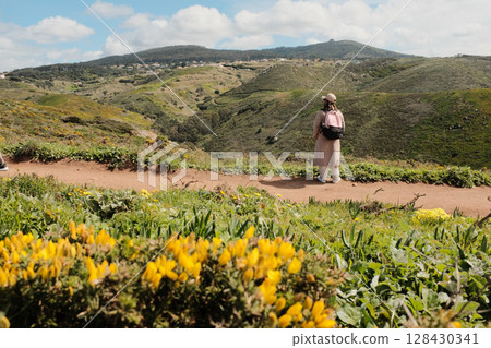 Woman enjoying springtime flower field on scenic hiking trail with mountain hills in background at Cabo da Roca Sintra Portugal nature walk with wild yellow blooms and blue sky. High quality photo 128430341