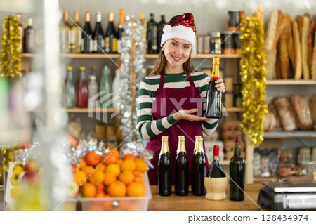 Friendly young girl shop seller posing behind counter with alcoholic drinks, Christmas goods 128434974