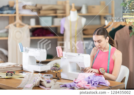 Woman seamstress sews on sewing machine in workshop Woman seamstress sews on sewing machine in workshop 128435002