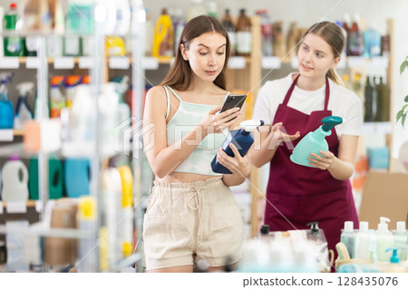 Female shopper scans QR code on label of spray glass cleaner using her smartphone. Young sales girl helps to choose spray glass cleaner in supermarket 128435076
