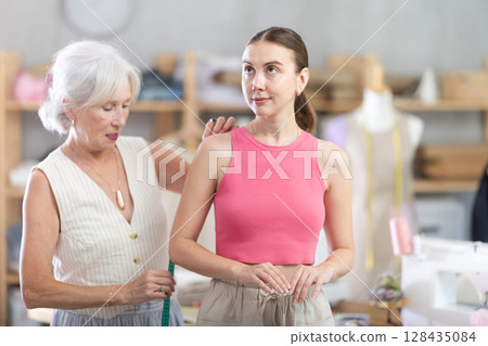 Senior woman tailor measures female client Senior woman tailor measures female client 128435084