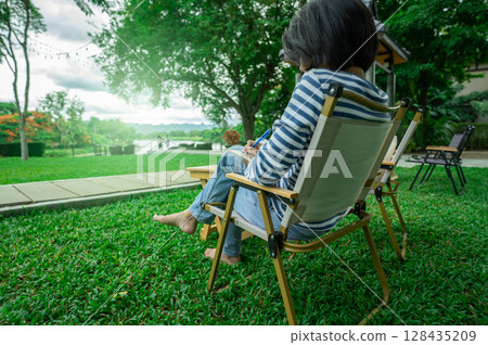 Woman enjoying digital detox while reading book in peaceful garden chair surrounded by green nature for mental wellness and self-reflection time. Social media break. Mindful living and disconnection. 128435209