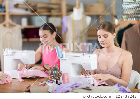 Adult and young women seamstresses sewing on machine Adult and young women seamstresses sewing on machine 128435242