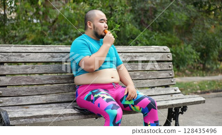 Fat yoga instructor munching on a carrot on a park bench 128435496