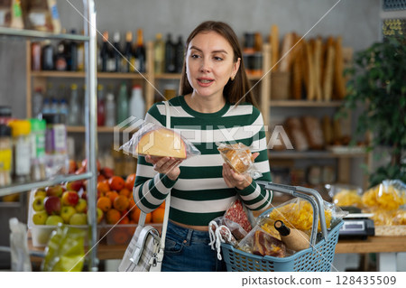 Smiling young girl with shopping basket trying to make choice holding two sorts of cheese in grocery store 128435509