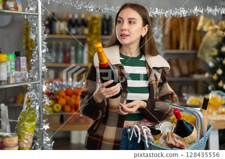 Interested young girl shopper holding bottle and choosing wine for holiday table in grocery hypermarket Interested young girl shopper holding bottle and choosing wine for holiday table in grocery hypermarket 128435556