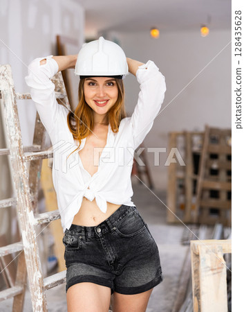 Brown-haired woman in construction hardhat, shorts and unbuttoned blouse standing in apartment during overhaul 128435628