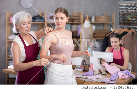 Senior female tailor take measurements from girl client, adult woman worker sewing in background 128435655
