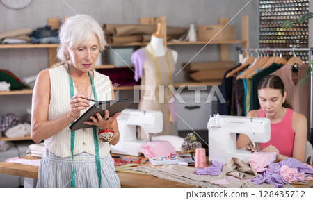 Adult seamstress sewing woman assistant drawing pattern on tablet Adult seamstress sewing woman assistant drawing pattern on tablet 128435712