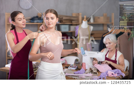 Female tailor take measurements from girl client, mature woman worker sewing in background 128435725