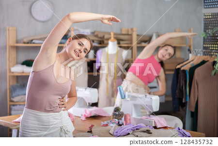 Young and adult women doing warm-up exercises in sewing workshop Young and adult women doing warm-up exercises in sewing workshop 128435732