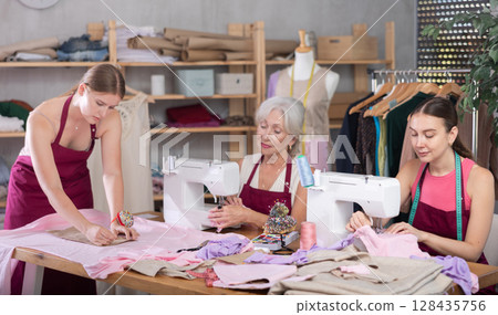 Two craftsperson sew clothes, girl assistant attaches pattern to fabric. 128435756