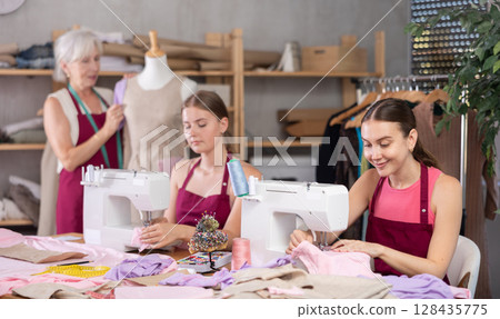 Two seamstresses sewing next to an elderly dressmaker in an atelier Two seamstresses sewing next to an elderly dressmaker in an atelier 128435775