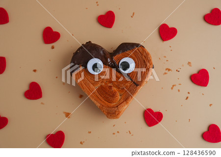 Funny creative eyes on Heart shaped dark chocolate croissant on beige background with copy space. Puff pastry new trendy sweet dessert bakery. Creamy dough Funny creative eyes on Heart shaped dark chocolate croissant on beige background with copy space. Puff pastry new trendy sweet dessert bakery. Creamy dough 128436590