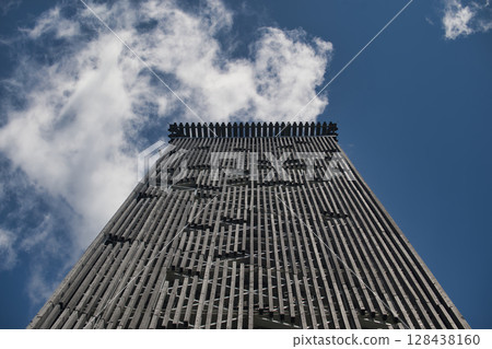 A modern wooden tower reaching towards a vibrant blue sky with soft white clouds. 128438160