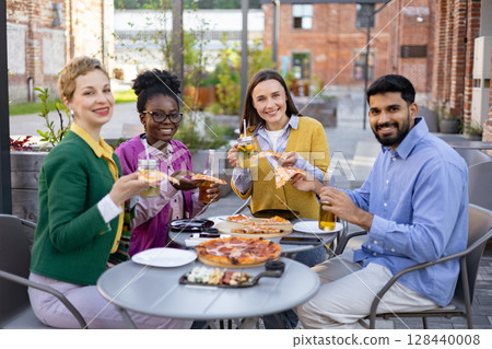 Diverse group of friends enjoying pizza and drinks together outdoors at a restaurant. 128440008