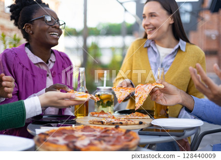 Friends sharing pizza outdoors, laughing and enjoying each other's company at a table. 128440009