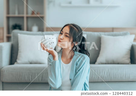Young Asian woman holding piggy bank and sitting on the floor at home 128440312
