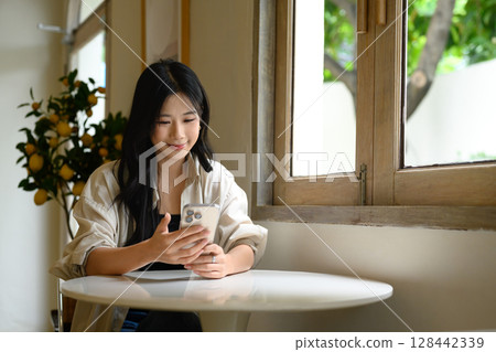 Asian woman checking her phone while enjoying a relaxing time at a modern cafe Asian woman checking her phone while enjoying a relaxing time at a modern cafe 128442339