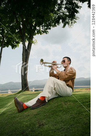 Musician sitting on a grassy hill playing trumpet under a tree with mountain scenery in the background Musician sitting on a grassy hill playing trumpet under a tree with mountain scenery in the background 128442369