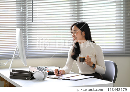 Young businesswoman working at her desk while holding a cup of coffee during a productive day Young businesswoman working at her desk while holding a cup of coffee during a productive day 128442379