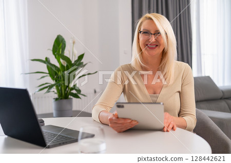 Portrait of cheerful female therapist in cozy setting conducting remote consultation using laptop and tablet. Modern online mental health care. 128442621