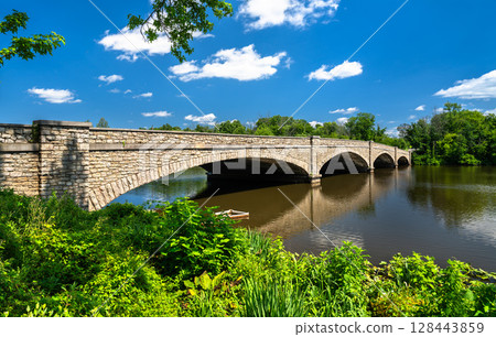 Stone-arched Washington Road Bridge spanning Carnegie Lake in Princeton, New Jersey, United States. The historic structure is surrounded by lush greenery and reflected in the calm lake water beneath a Stone-arched Washington Road Bridge spanning Carnegie Lake in Princeton, New Jersey, United States. The historic structure is surrounded by lush greenery and reflected in the calm lake water beneath a 128443859