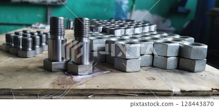 Close-up of new metal bolts and nuts. Steel bolts and nuts. Industrial fasteners displayed on a wooden surface, with a blurred workshop background. Flanged steel hex nuts. Mechanical engineering. 128443870