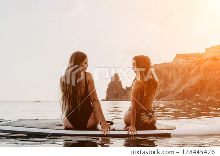 Women Paddleboard Beach Sunset - Two women sit on a paddleboard in the ocean at sunset, looking at each other. 128445426