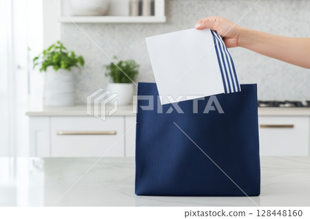 A hand is inserting a folded white envelope into a stylish navy blue tote bag resting on a clean kitchen 128448160