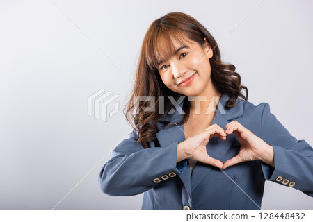 A confident business woman wear suit smiles and creates a heart symbol with two hands on white background. Asian portrait of a beautiful young female expressing love and happiness for Valentine's Day 128448432