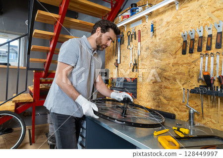 Skilled bike mechanic man using tools repairing bicycle in well-organized workshop Skilled bike mechanic man using tools repairing bicycle in well-organized workshop 128449997