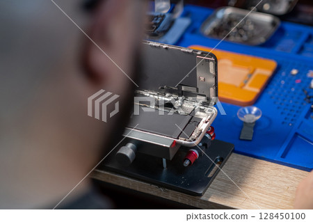 A technician meticulously replaces a smartphone battery in a well-equipped workshop with various tools nearby. A technician meticulously replaces a smartphone battery in a well-equipped workshop with various tools nearby. 128450100