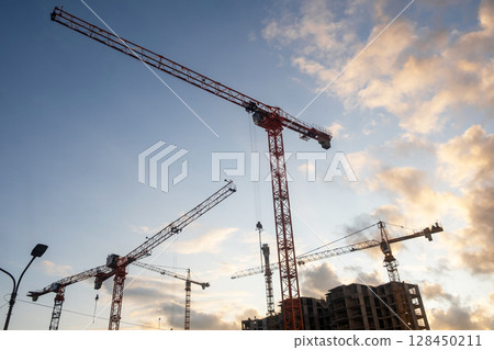 Cranes are actively lifting materials at a construction site, silhouetted by a colorful sunset in the background. Cranes are actively lifting materials at a construction site, silhouetted by a colorful sunset in the background. 128450211