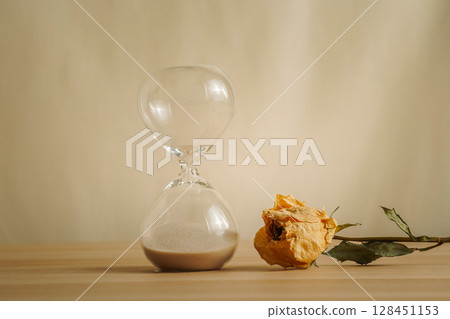 A simple hourglass on a wooden table with dry rose a beige backdrop, symbolizing time flow, aging, and minimalistic living. Represents timeless themes in a creative presentation for stylish concepts. A simple hourglass on a wooden table with dry rose a beige backdrop, symbolizing time flow, aging, and minimalistic living. Represents timeless themes in a creative presentation for stylish concepts. 128451153